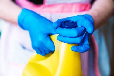 Person with blue latex gloves and apron opening a yellow bottle of bleach