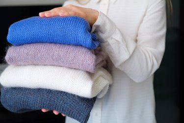A woman or a girl holds an armful of knitted things of different colors, stacked in a pile, in the room. Winter and autumn warm cozy sweaters for charity. The concept of storage, care and washing of handmade products. A copy of the text space.