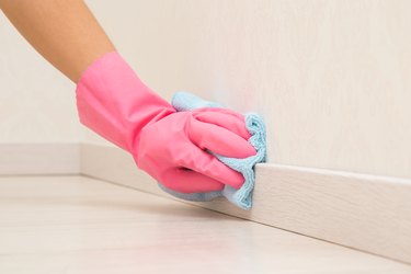 Hand in pink rubber protective glove using blue dry rag and wiping light wooden baseboard surface in room at home